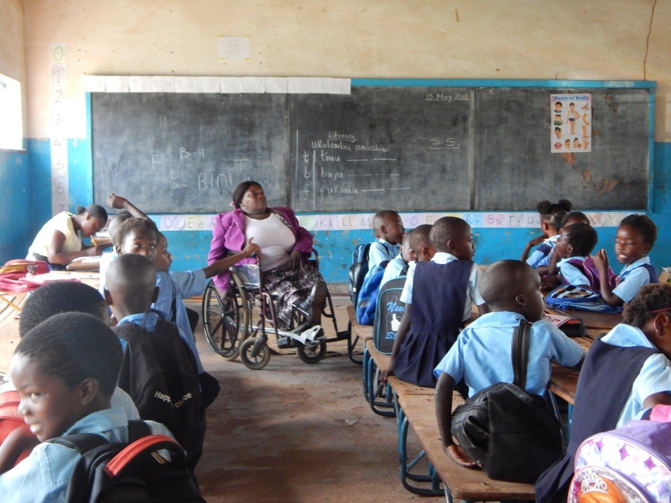 Sambialaisessa luokkahuoneessa pyörätuolissa istuva opettaja opettaa ja lapset istuvat reput selässä pulpeteissaan