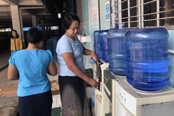 Kaksi tehtaan työntekijänaista myanmarilaisen vaatetahtaan vedenjakelupisteellä. Kolmas nainen katselee kauempaa.