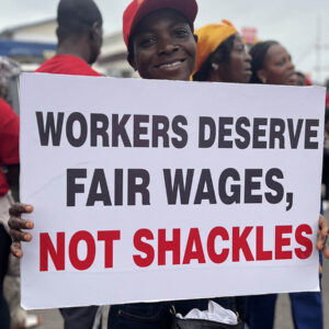 Afrikkalainen nainen pitää mielenosoituksessa kylttiä, jossa lukee: Workers deserve fair wages, not shackles