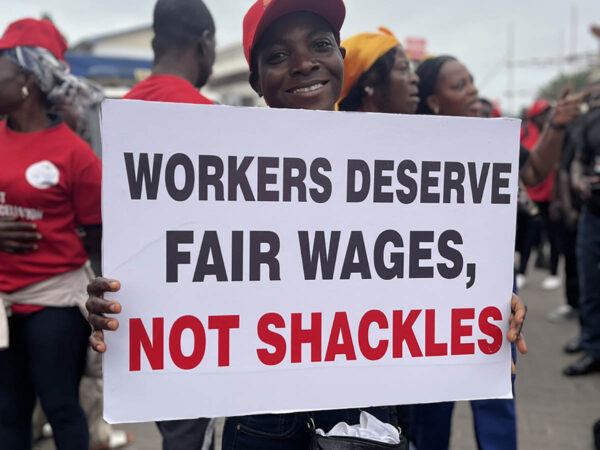Afrikkalainen nainen pitää mielenosoituksessa kylttiä, jossa lukee: Workers deserve fair wages, not shackles
