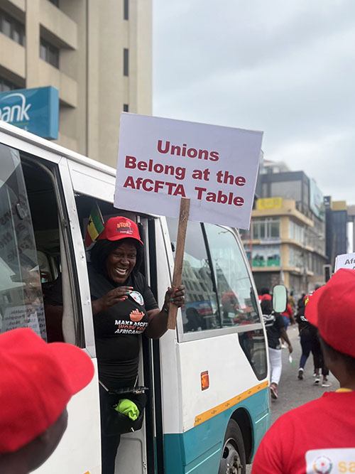 Mielenosoittajanainen Ghanassa kurkistaa bussista kädessään kyltti: Unions Belong at the AfCFTA Table